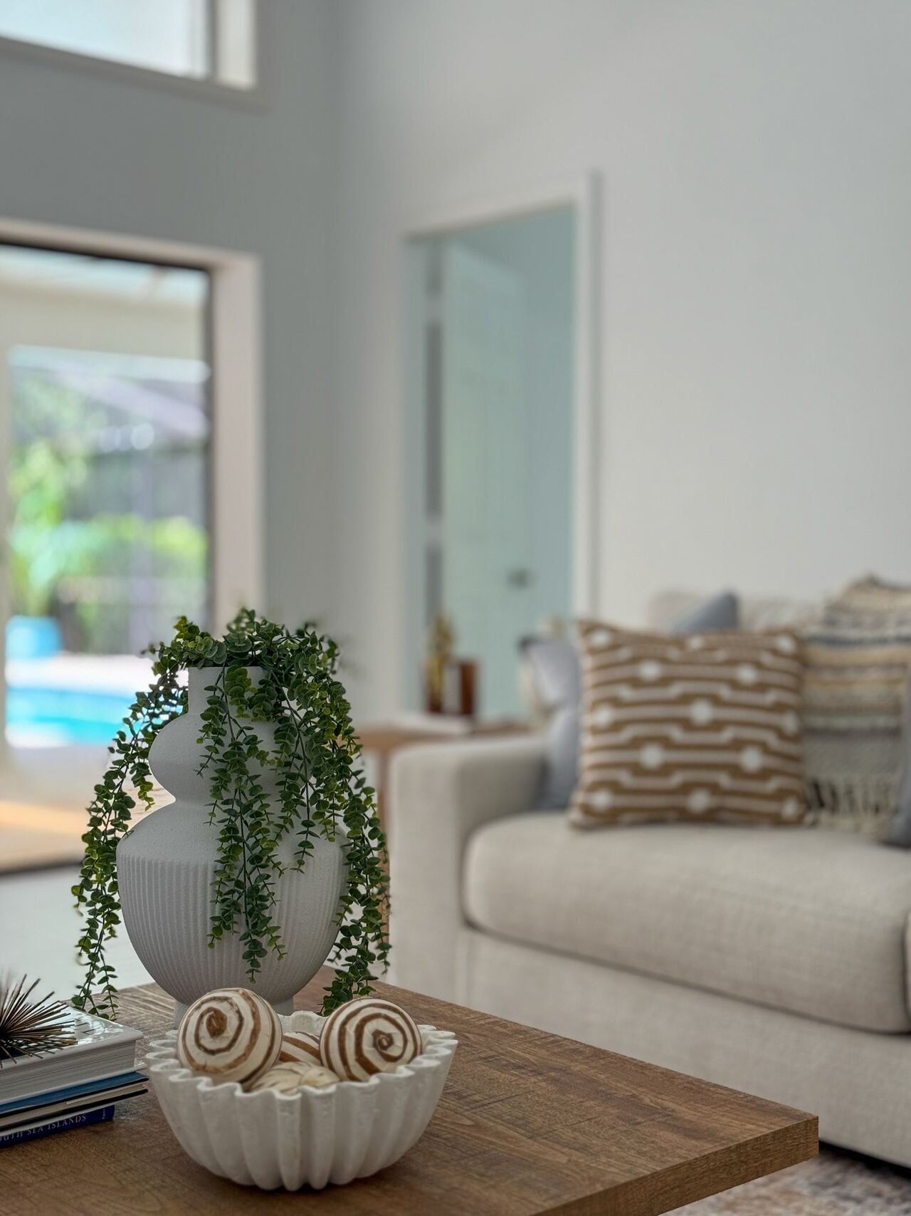 Close-up of a white ribbed vase with trailing greenery beside a scalloped bowl of shells on a wood coffee table, with the pool visible behind