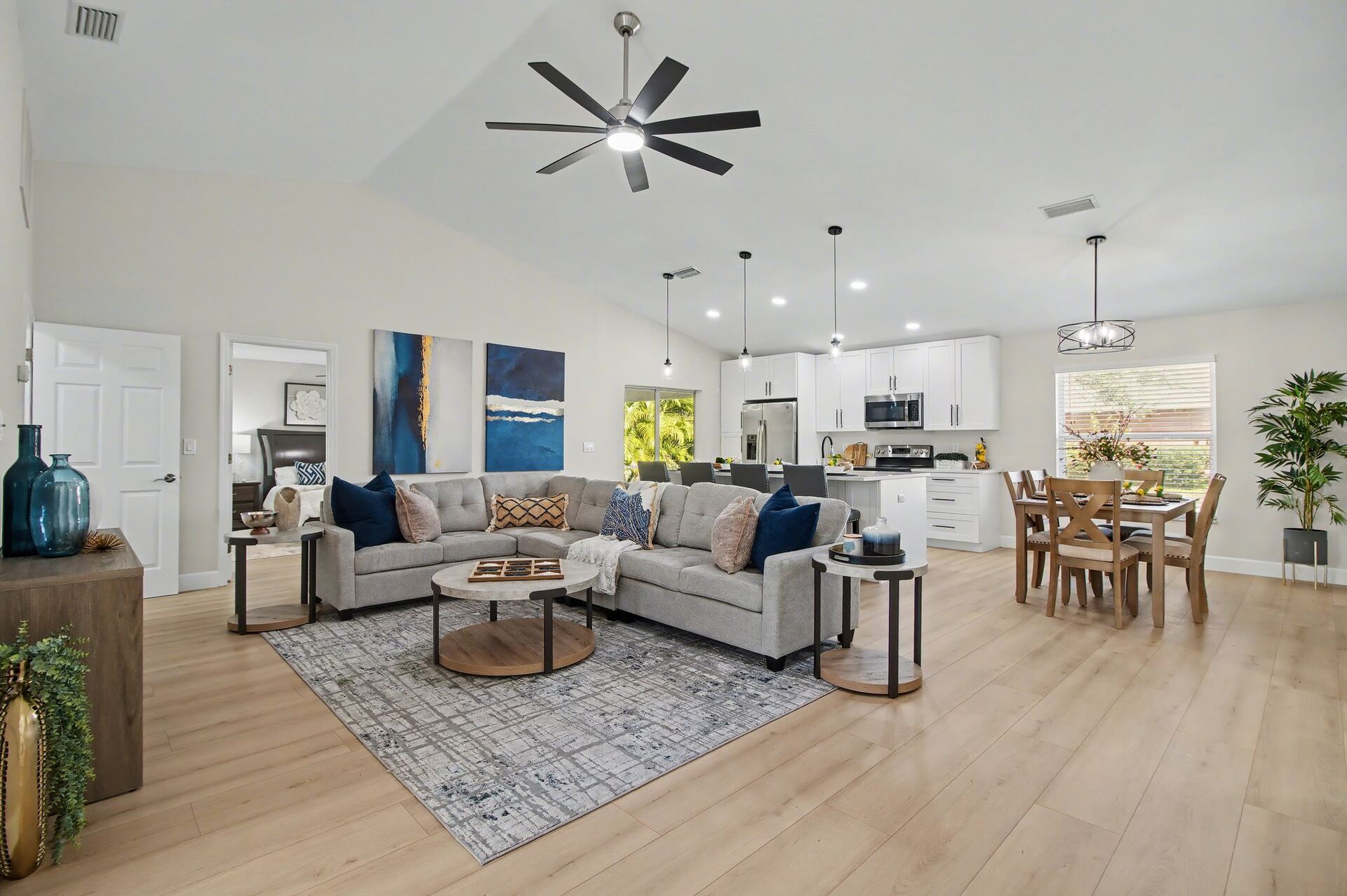 Open-concept staged great room flowing into a white kitchen and dining area, with a vaulted ceiling, large dark ceiling fan, gray sectional and navy and gold diptych artwork