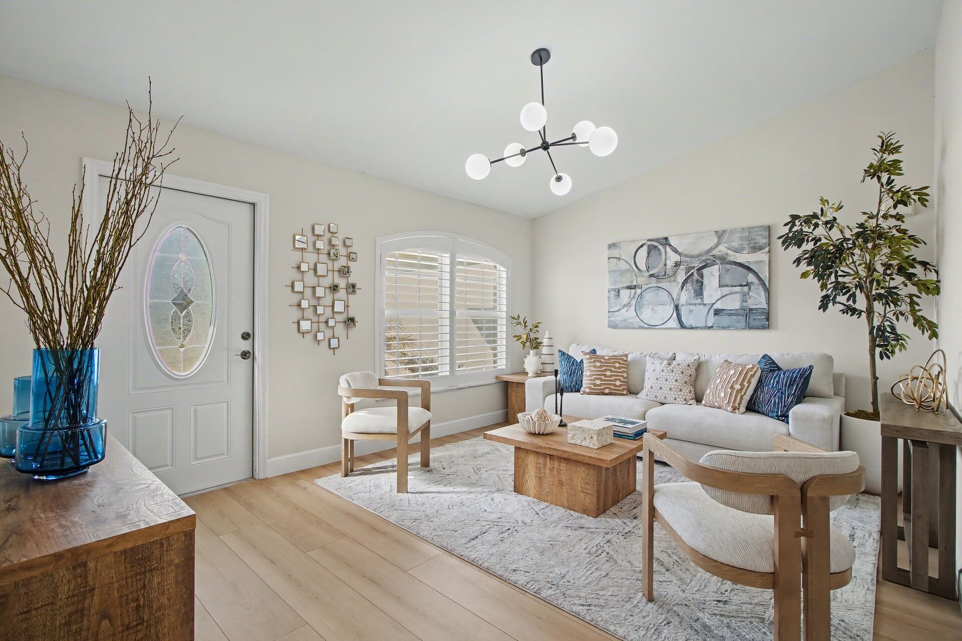 Staged living room with blue glass vases on a wood credenza, an oval leaded-glass front door, gold mirrored wall art and a black sputnik chandelier