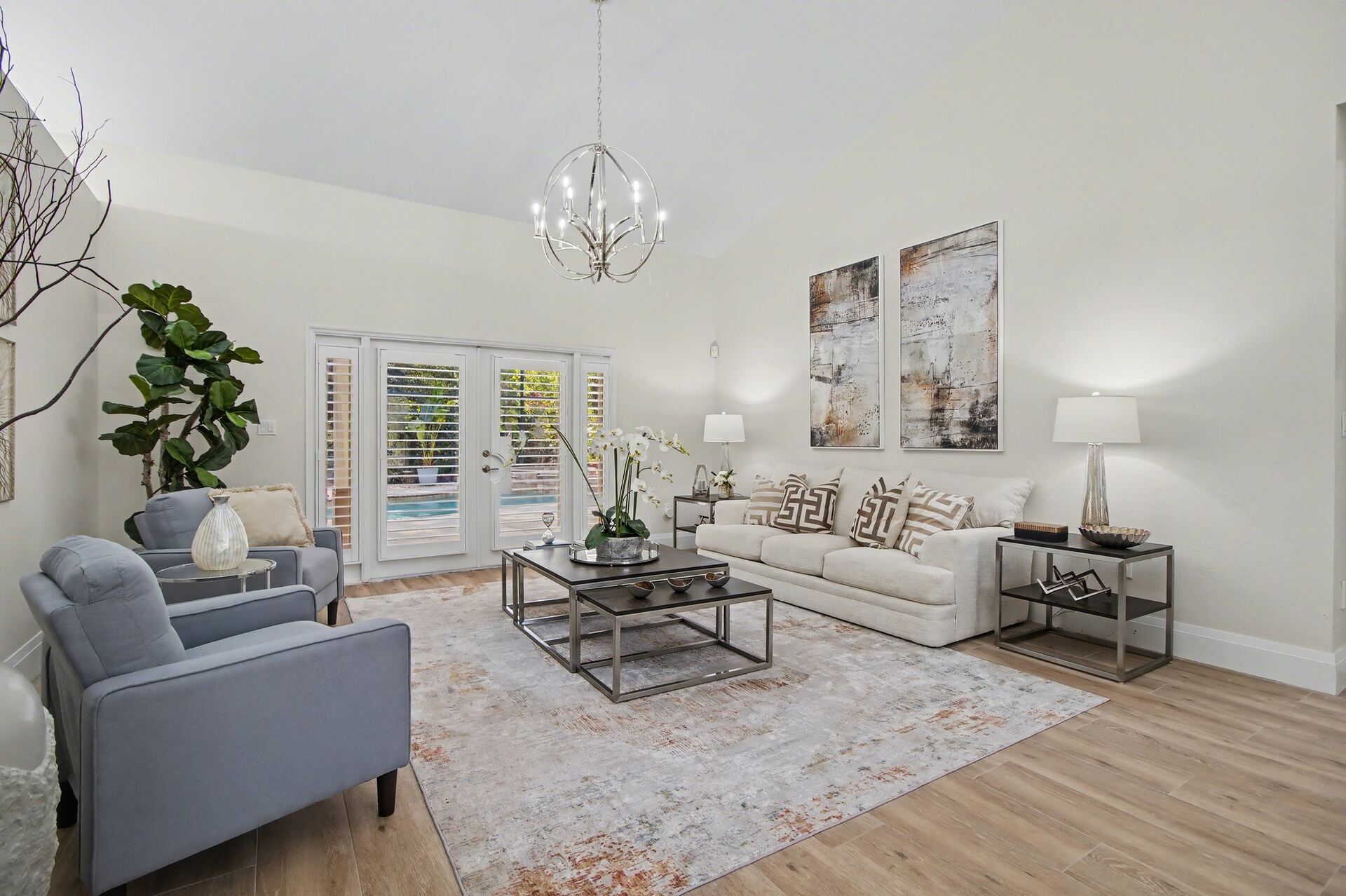 The formal staged living room from the opposite angle, with an orb chandelier, blue armchairs and french doors to the pool