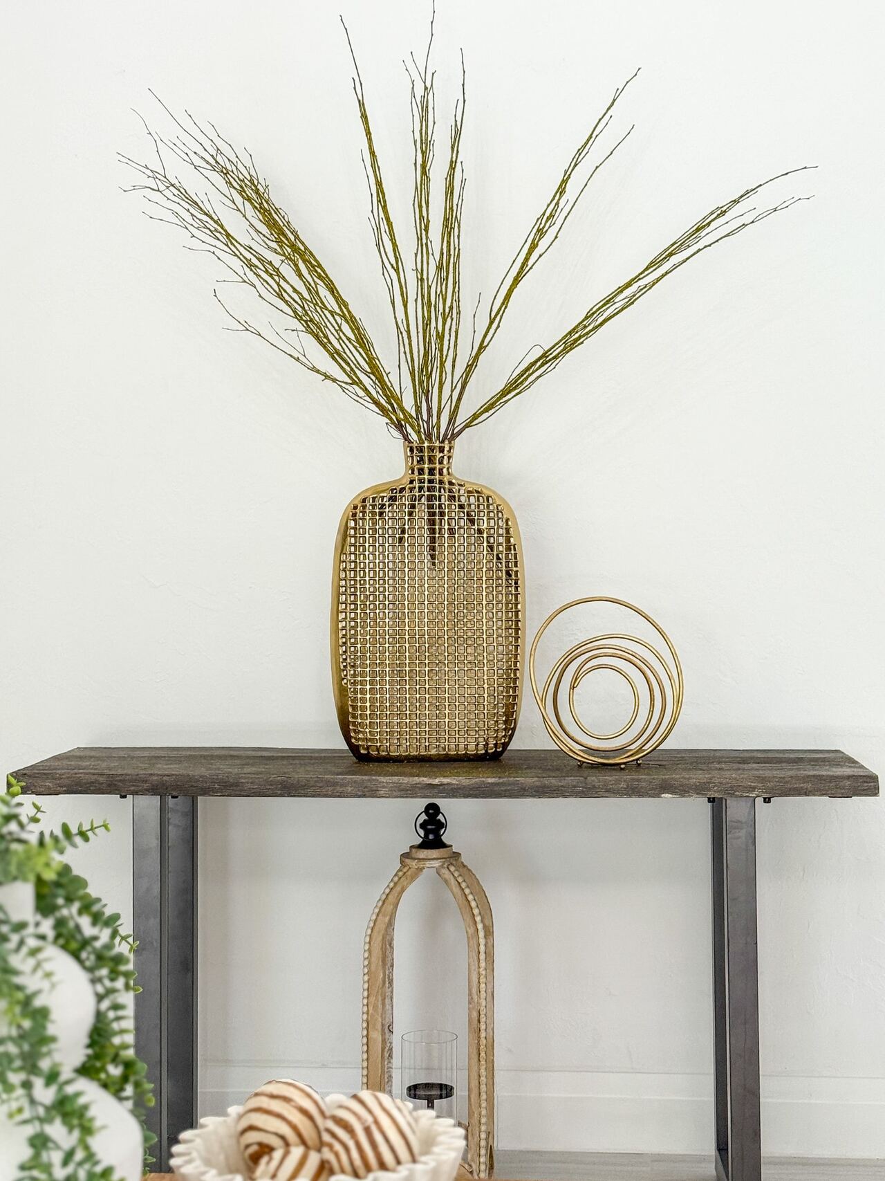 Tall detail of a gold woven vase holding tall grass branches on a gray console table, with a gold spiral sculpture beside it