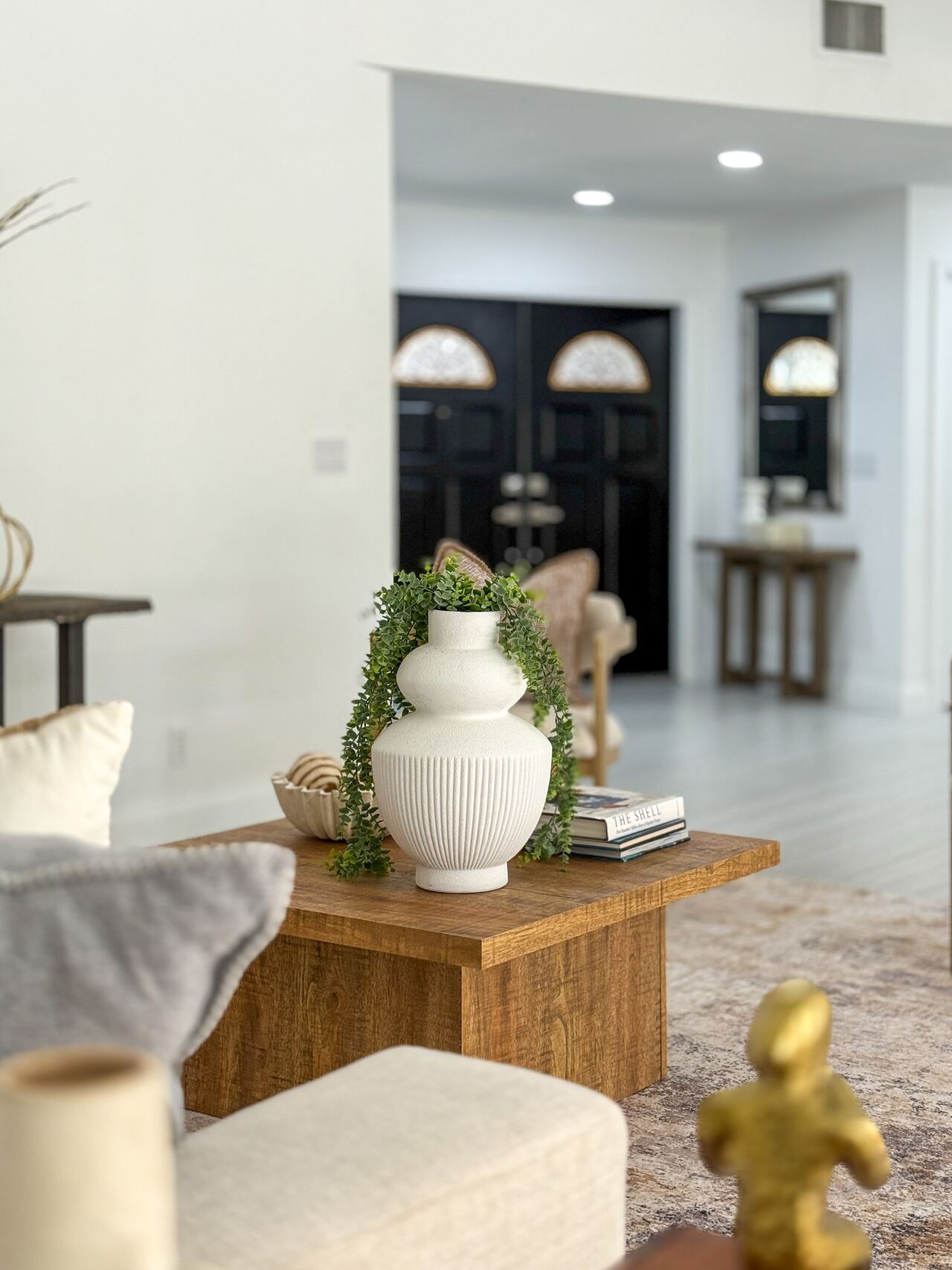Coffee table vignette with a white ribbed vase and trailing greenery, with black double front doors far in the background