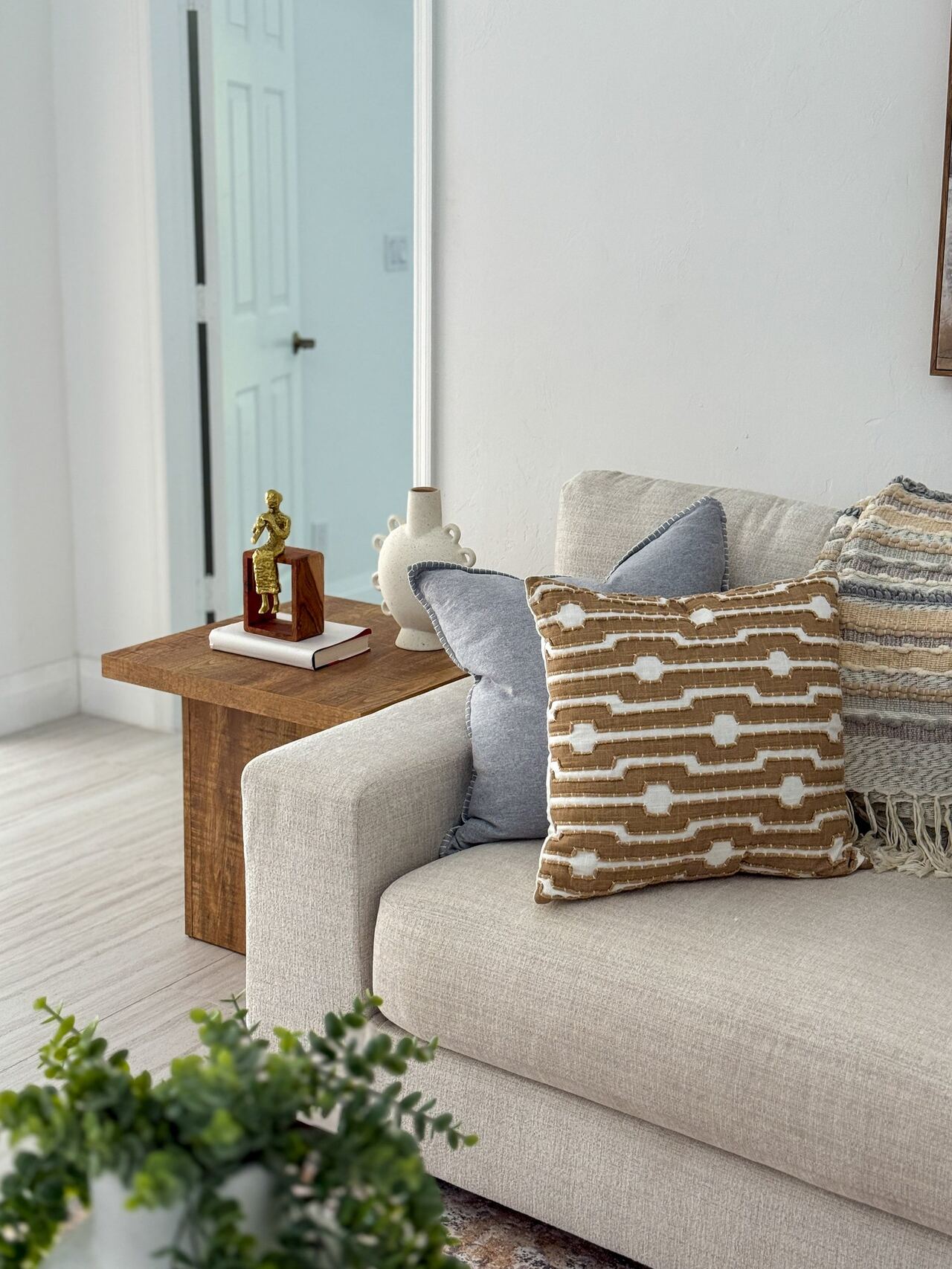 Sofa arm beside a wood side table holding a gold figurine on stacked books, with tan and blue pillows
