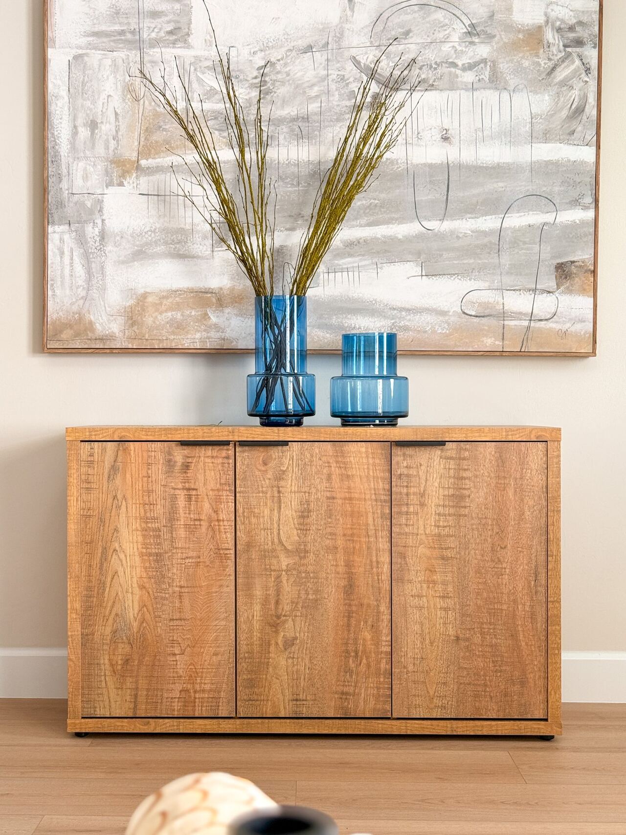 Wood three-door credenza topped with two blue glass vases and branches beneath a large abstract canvas