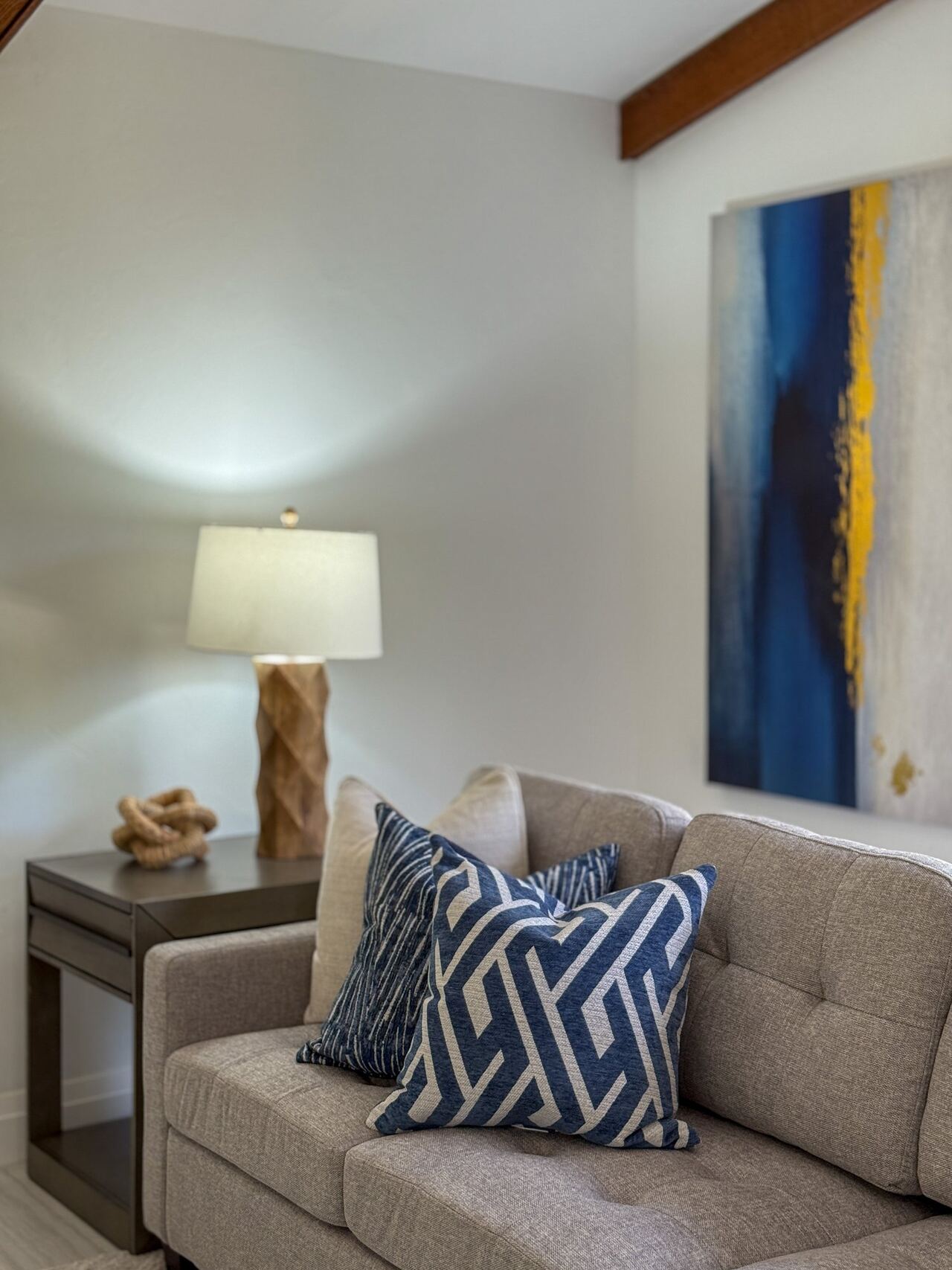 Tufted sofa styled with blue geometric pillows beneath a blue and yellow abstract canvas, under a wood ceiling beam