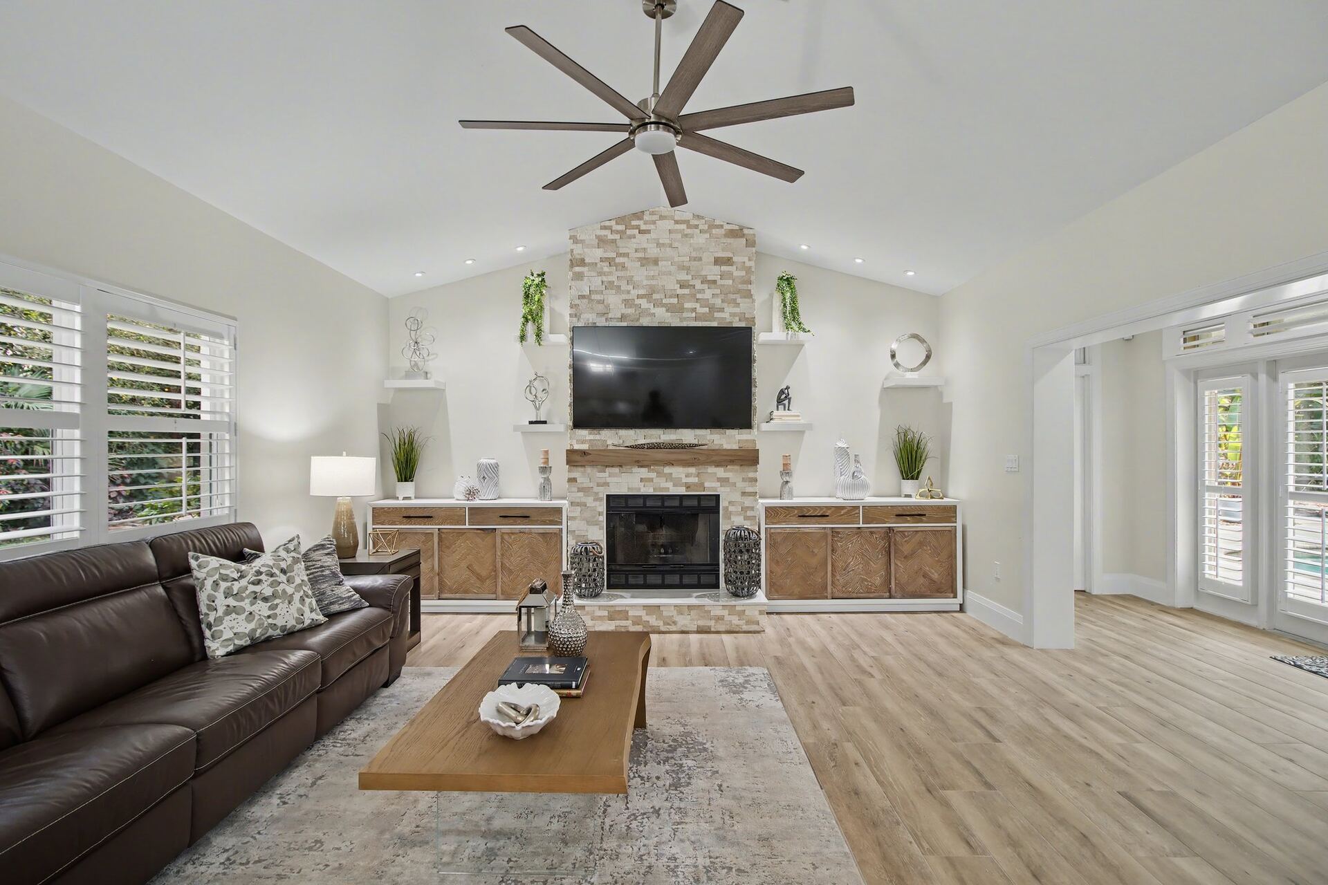 Staged great room with a stacked-stone fireplace, mounted TV, large wood-blade ceiling fan and a brown leather sofa