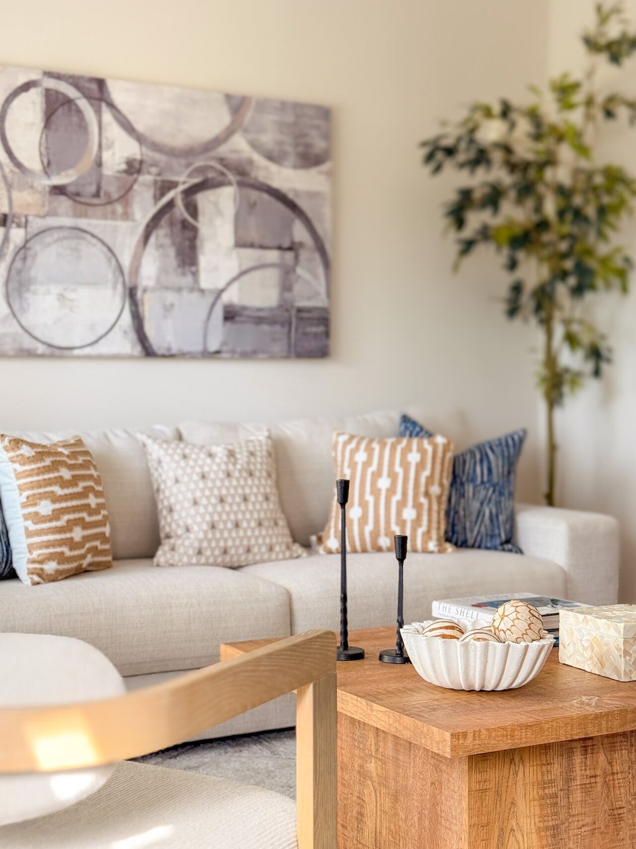 Cream sofa with tan and blue pillows beneath a circular abstract canvas, with black candlesticks on a wood coffee table